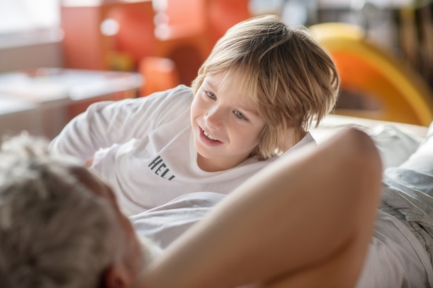 Comprensione. Bambino sorridente credulone in camicetta bianca che comunica con papà che è sdraiato a casa sul letto con le spalle alla telecamera