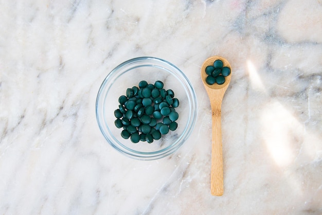 compresse di spirulina in una ciotola di vetro e un cucchiaio di legno su un vassoio di legno su uno sfondo di marmo grigio