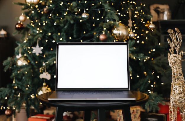 Computer portatile con schermo vuoto bianco mock up, sul tavolo vicino all'albero di Natale.