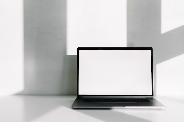 Computer portatile con schermo vuoto bianco sul tavolo bianco con ombra dalla finestra.