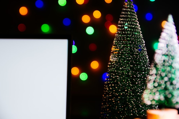Computer portatile con schermo vuoto su tavolo di legno, albero di Natale sfocato e regali sullo sfondo. Carta di Natale e felice anno nuovo, bokeh, scintille. Disposizione piatta, vista dall'alto