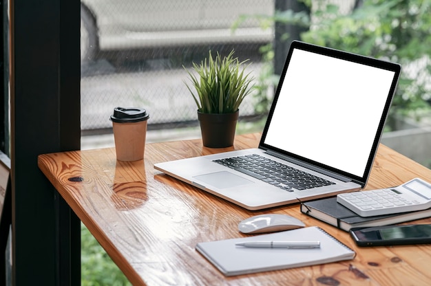 Computer portatile e forniture dello schermo in bianco del modello sulla tavola di legno nella stanza dell'ufficio di stile del sottotetto.