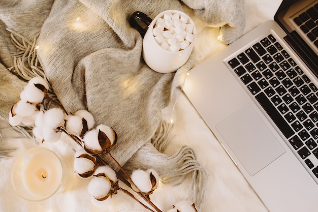 Computer portatile e tazza di cacao con marshmallow, sciarpa, fiori di cotone, luci scintillanti, candela sul letto bianco. Concetto di lavoro a casa. Autunno, autunno, inverno concetto. Lay piatto, vista dall'alto, copia spazio.