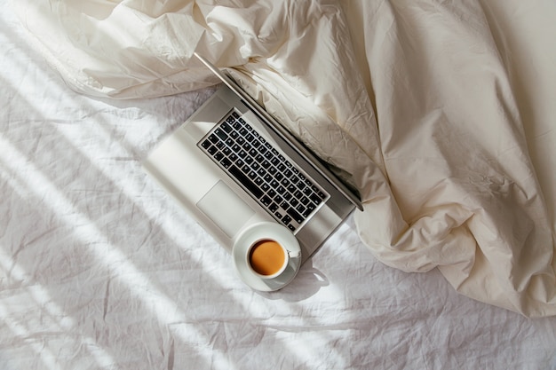 Computer portatile e tazza di caffè sul letto bianco con una coperta. Concetto di lavoro a casa. Luce del mattino