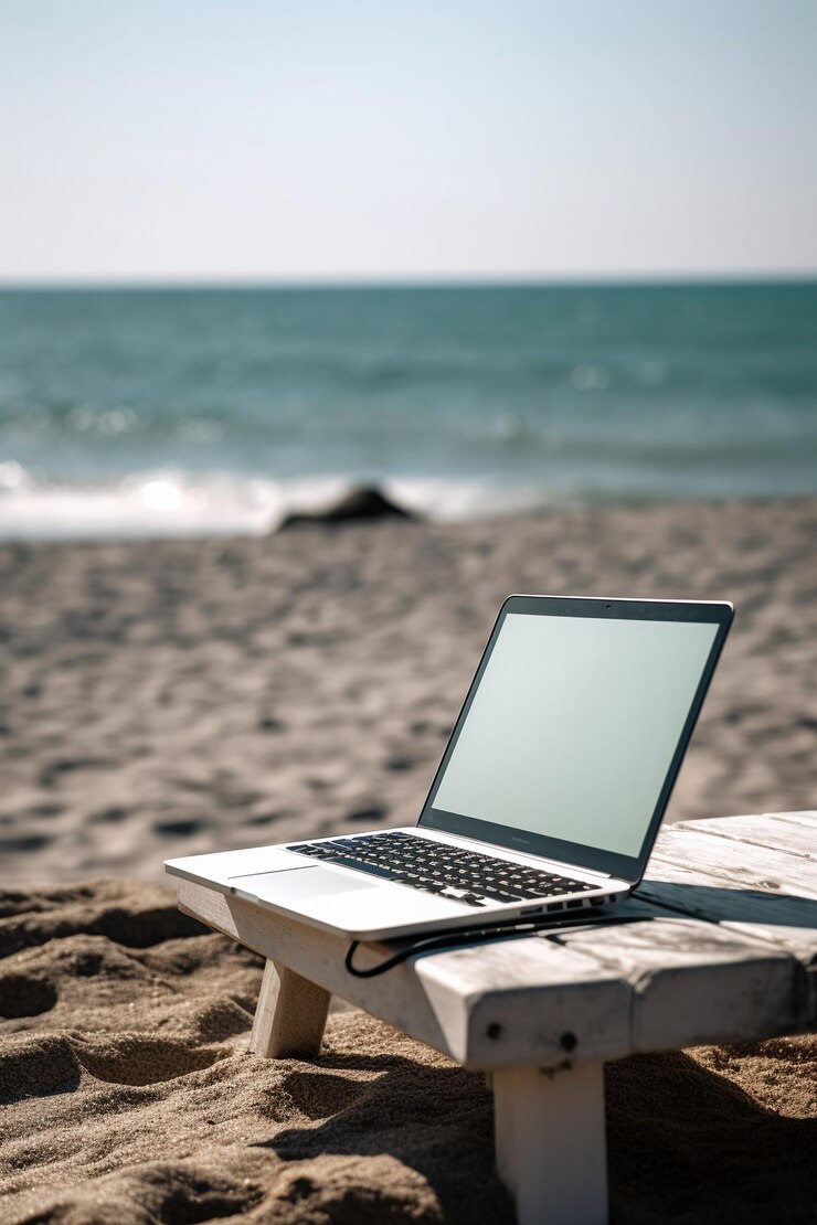 Computer portatile su una panca di legno su una spiaggia con l'oceano sullo sfondo.