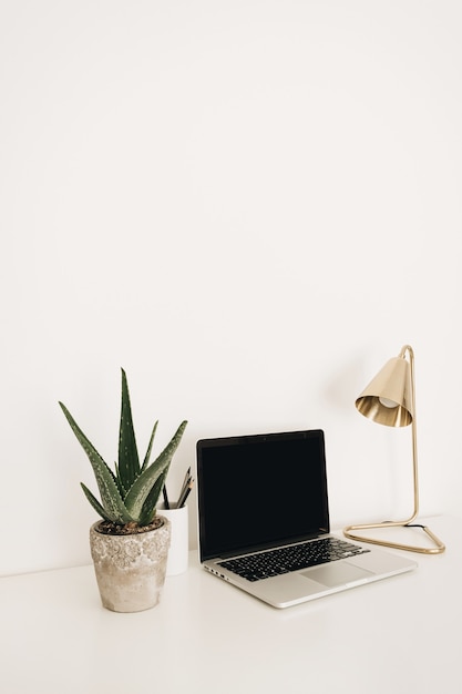 Computer portatile sul tavolo bianco con lampada dorata e pianta di aloe. Area di lavoro minimale della scrivania dell'home office con modello di mockup