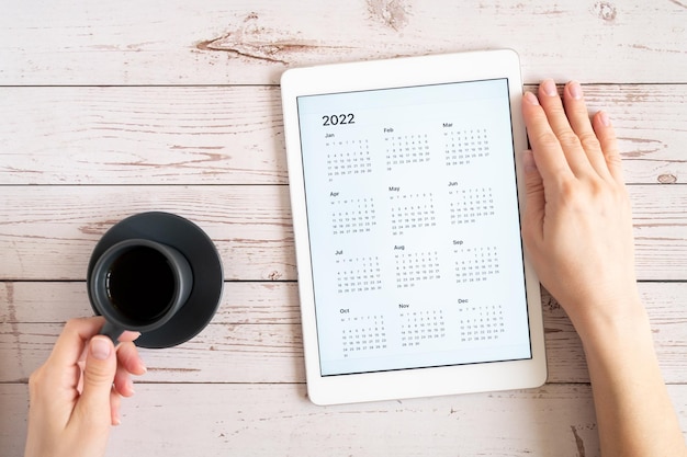 Computer tablet con un'app aperta del calendario per l'anno 2022 e tazza di tè o caffè nelle mani della donna su fondo di legno. concetto di business o per elencare gli obiettivi con l'utilizzo della tecnologia. vista dall'alto, posizione piatta