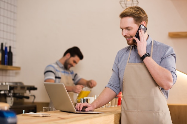 Comunicazione a distanza. Uomo piacevole gioioso positivo guardando lo schermo del laptop e parlando al telefono mentre si lavora sul laptop