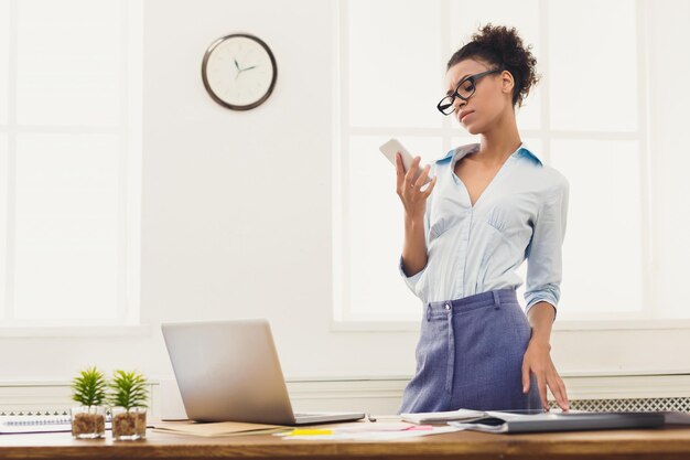 Comunicazione, tecnologia, successo. Donna d'affari afroamericana che utilizza il telefono cellulare, in piedi sul posto di lavoro in ufficio, copia spazio