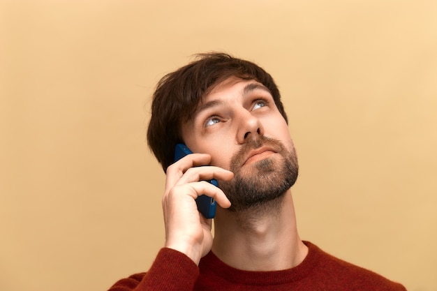 Comunicazioni. La foto del giovane con la barba ha una conversazione telefonica, tiene il cellulare vicino all'orecchio, dice notizie ad un amico, si concentra, indossa un maglione, posa contro il muro beige