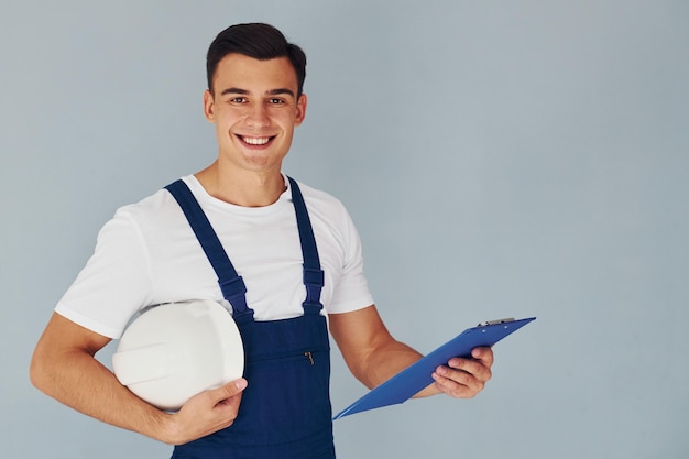 Con blocco note e elmetto Lavoratore maschio in uniforme blu in piedi all'interno dello studio su sfondo bianco