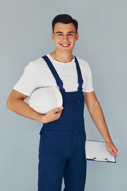Con blocco note e elmetto Lavoratore maschio in uniforme blu in piedi all'interno dello studio su sfondo bianco