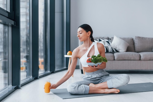 Con cibo sano e succo d'arancia in mano Giovane donna con una forma del corpo snella in abbigliamento sportivo ha una giornata di fitness al chiuso a casa