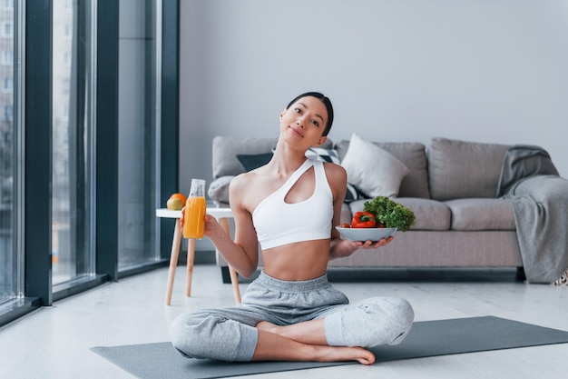 Con cibo sano e succo d'arancia in mano Giovane donna con una forma del corpo snella in abbigliamento sportivo ha una giornata di fitness al chiuso a casa