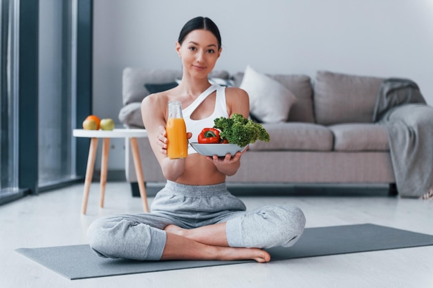 Con cibo sano e succo d'arancia in mano Giovane donna con una forma del corpo snella in abbigliamento sportivo ha una giornata di fitness al chiuso a casa