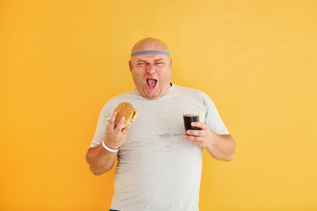 Con il fast food Un uomo in sovrappeso divertente con una cravatta sportiva sul fondo giallo