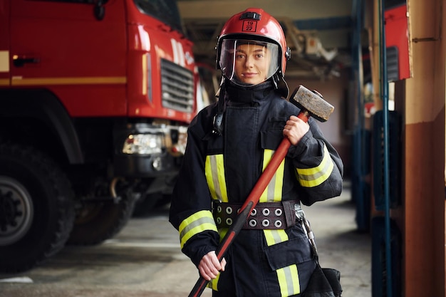 Con il martello in mano Vigile del fuoco femminile in uniforme protettiva in piedi vicino al camion