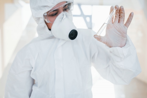 Con la provetta in mano. Scienziato medico femminile in camice da laboratorio, occhiali difensivi e maschera in piedi al chiuso