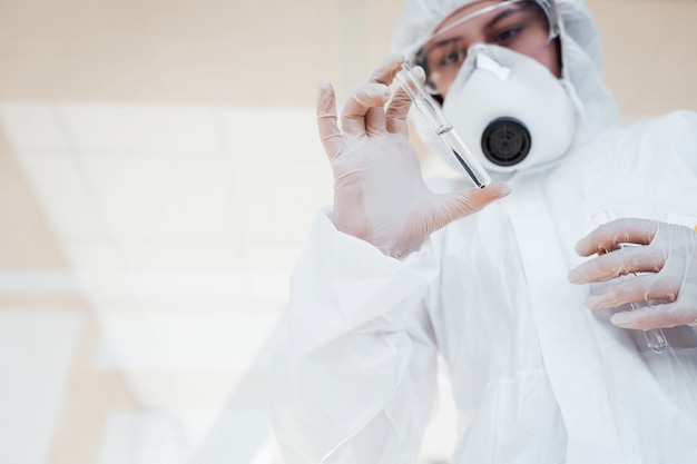 Con la provetta in mano. Scienziato medico femminile in camice da laboratorio, occhiali difensivi e maschera in piedi al chiuso