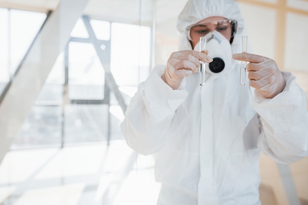 Con la provetta in mano. Scienziato medico femminile in camice da laboratorio, occhiali difensivi e maschera in piedi al chiuso