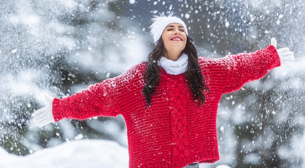 Con le braccia spalancate, la donna in maglione rosso, guanti bianchi, sciarpa e cappello gode di aria fresca in una giornata invernale innevata.