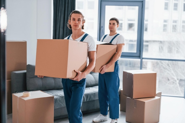 Con le scatole in mano Due giovani traslocatori in uniforme blu che lavorano al chiuso nella stanza