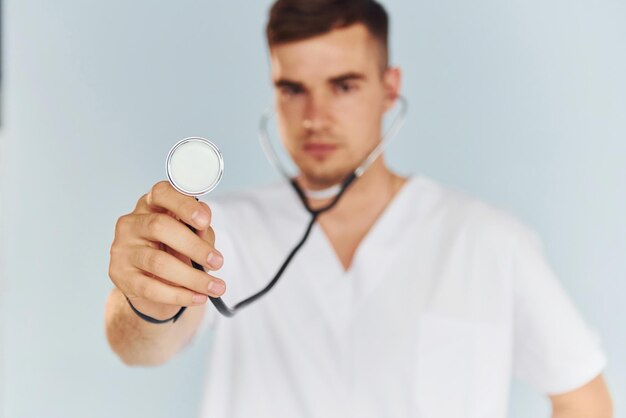 Con lo stetoscopio Giovane medico maschio in uniforme è al chiuso su sfondo bianco