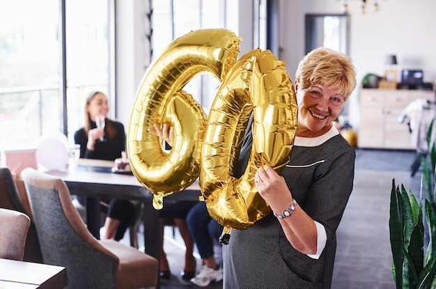 Con palloncini del numero 60 in mano. Senior donna con la famiglia e gli amici per festeggiare un compleanno al chiuso.