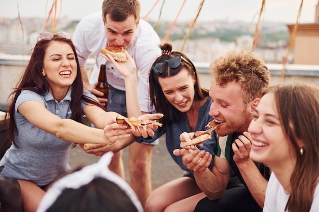 Con una deliziosa pizza Un gruppo di giovani in abiti casual organizzano una festa sul tetto insieme durante il giorno