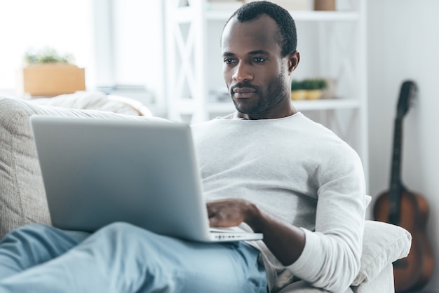 Concentrandosi su qualcosa. Bel giovane africano che guarda il laptop con una faccia seria mentre è sdraiato sul divano di casa