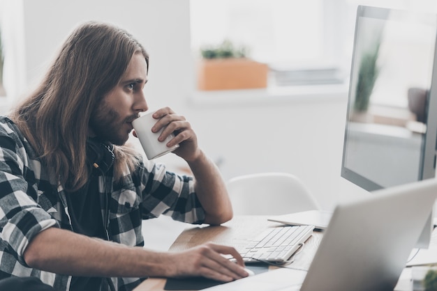 Concentrarsi sul lavoro. Bel giovane con i capelli lunghi che lavora al computer e beve caffè mentre è seduto alla sua scrivania in un ufficio creativo