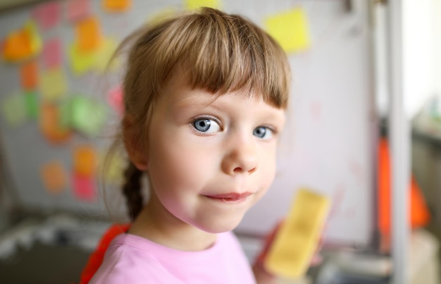 Concentrarsi sulla bambina che guarda la fotocamera con uno sguardo strano e illeggibile sul viso carino e in piedi in un'aula moderna con una tavola di vetro hightech Sfondo sfocato