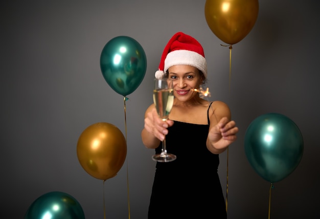 Concentrati su una bella donna africana con cappello da Babbo Natale e abito nero che celebra la festa di Capodanno con stelle filanti e flauto di champagne in mano contro mongolfiere lucide su sfondo grigio muro con spazio pubblicitario copia