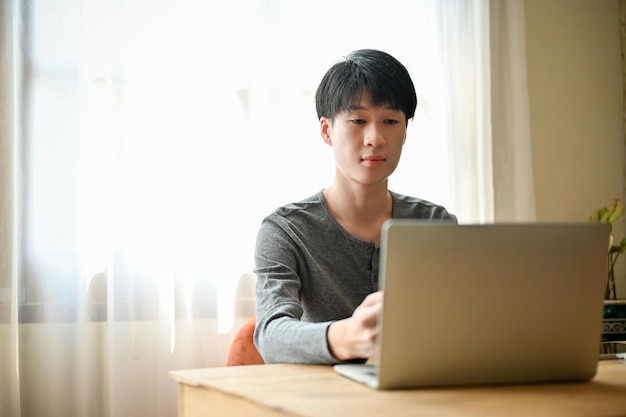 Concentrato giovane uomo asiatico utilizzando il computer portatile a una scrivania nel suo salotto di casa