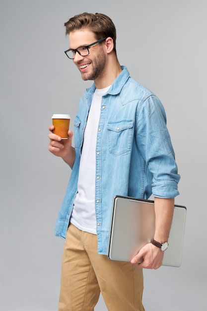 Concentrato giovane uomo barbuto con gli occhiali vestito in camicia di jeans che tiene laptop e tazza di caffè per andare