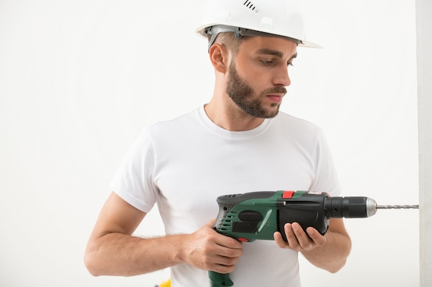 Concentrato giovane uomo barbuto in costruzione hardhat foratura nel muro durante la ristrutturazione nel nuovo appartamento