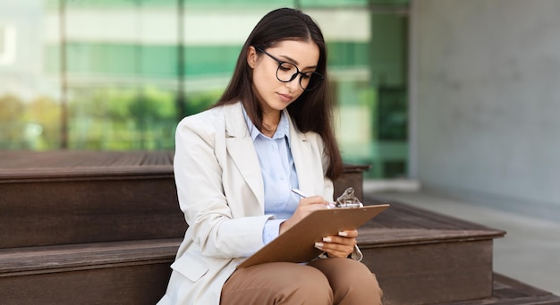 Concentrato serio bella giovane donna segretaria manager in abito occhiali scrivere a clipboard lavoro con