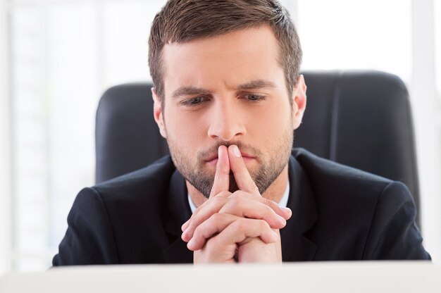 Concentrato sul lavoro. Bel giovane uomo formale guardando il computer portatile mentre è seduto al suo posto di lavoro