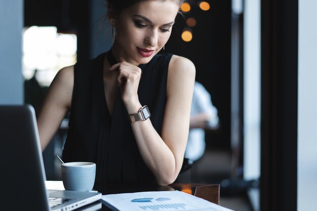 Concentrato sul lavoro. Fiduciosa giovane donna in abbigliamento casual intelligente che lavora al computer portatile mentre è seduta vicino alla finestra in un ufficio creativo o in un bar.