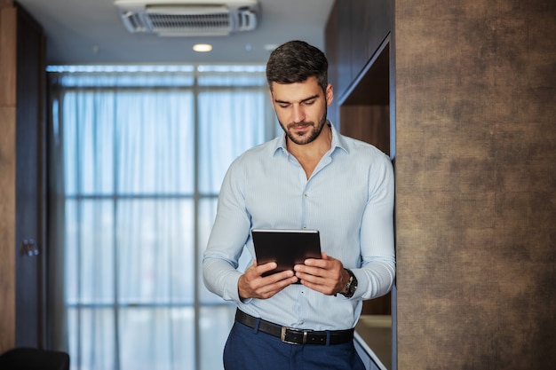 Concentrato sul lavoro. Un bel giovane formalmente e ben vestito che usa una tavoletta digitale mentre si trova su uno sfondo moderno al chiuso. Le mani maschili tengono un tablet, amo il mio lavoro