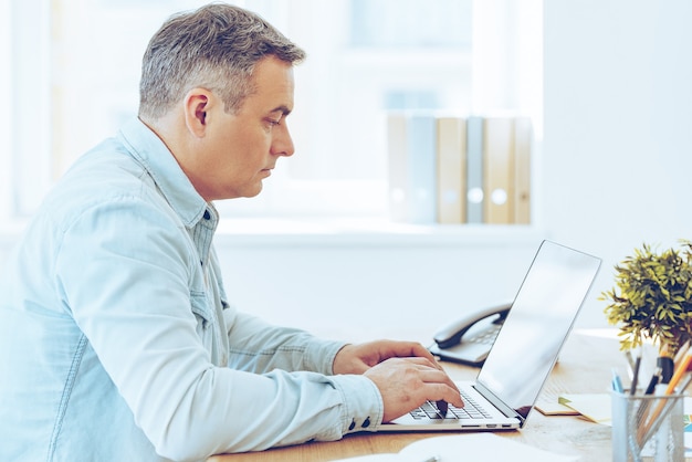 Concentrato sul lavoro. Vista laterale dell'uomo maturo che lavora al computer portatile mentre è seduto al suo posto di lavoro