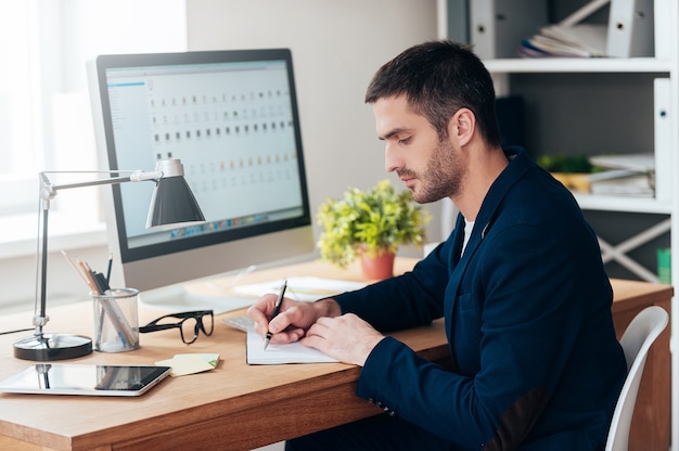 Concentrato sul lavoro. Vista laterale di un giovane fiducioso che scrive qualcosa sul suo taccuino mentre è seduto al suo posto di lavoro in ufficio