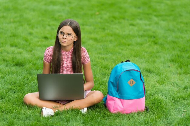 Concentrato sull'apprendimento del bambino con zaino e taccuino, nuova tecnologia nella vita moderna, il bambino serio lavora sul computer portatile l'istruzione online torna a scuola ragazza adolescente usa il computer sull'erba verde nel parco