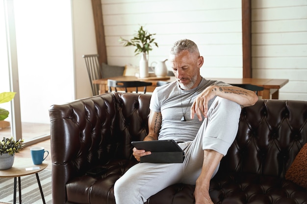 Concentrato uomo caucasico di mezza età in abiti casual utilizzando tablet digitale mentre è seduto sul divano in