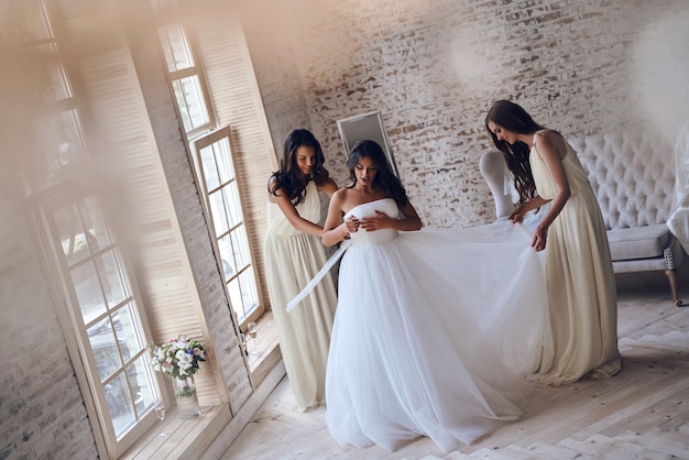 Concentrazione completa. Vista dall'alto a tutta lunghezza di due giovani donne attraenti che aggiustano un abito da sposa su una sposa mentre si trovano vicino alla finestra