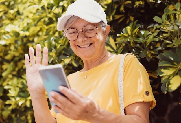 Concept di chiamata video Ritratto di una donna anziana sorridente in giallo seduta nel parco usando il telefono cellulare