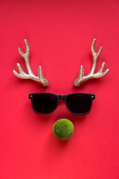 Concetto creativo del nuovo anno. Raindeer fatta di corna giocattolo scintillanti, sunglusses e palla verde su sfondo rosso. Biglietto di auguri festivo.