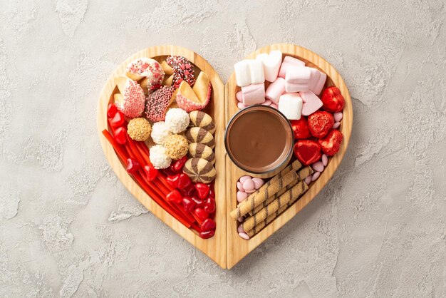 Concetto del giorno di San Valentino Top view foto di un vassoio in forma di cuore in legno con dolci, cioccolato, gelatina, biscotti e una tazza di vetro di cioccolato caldo sullo sfondo di texture di cemento