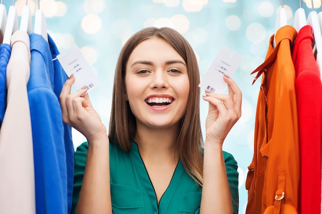 concetto di abbigliamento, moda, vendita, shopping e persone - donna felice che mostra i cartellini dei prezzi sui vestiti al guardaroba su sfondo blu luci vacanze