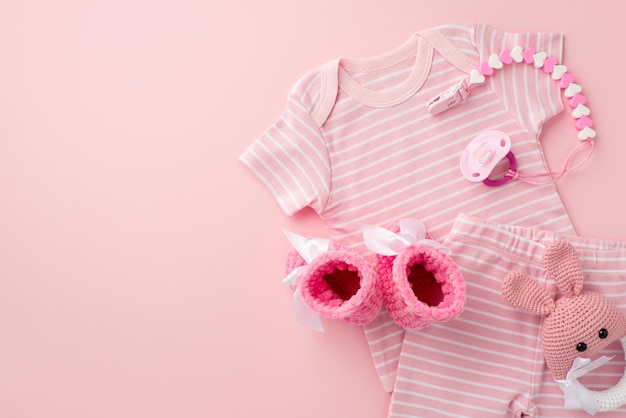 Concetto di accessori per bambini Foto vista dall'alto di vestiti per neonati rosa pantaloncini ciuccio catena stivaletti e giocattolo sonaglio coniglietto lavorato a maglia su sfondo rosa pastello isolato